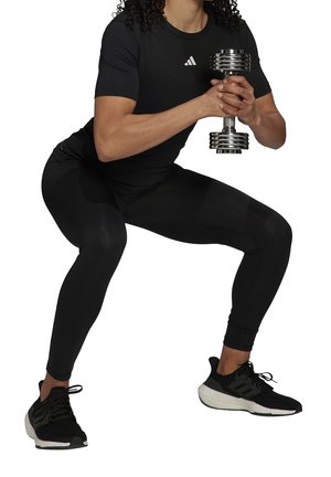 Osoba w czarnym stroju sportowym wykonująca przysiad, trzymająca srebrną hantlę obiema rękami.