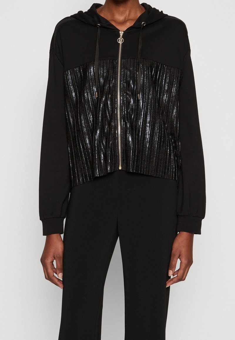 Sweat à capuche noir zippé avec un panneau avant plissé brillant, cordons à la capuche et manches longues, porté avec un pantalon noir.