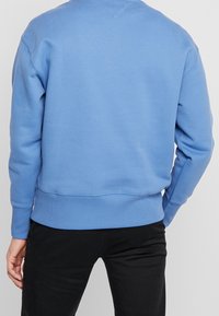 Un uomo con un maglione azzurro chiaro e pantaloni neri, visto di spalle, in piedi contro uno sfondo chiaro e uniforme.