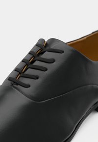 Black leather dress shoe with closed lacing and thin black laces, shown in close-up on a white background.