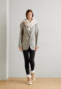 Blazer a cuadros con un corte entallado, bufanda beige de gran tamaño, mallas negras y botas marrones con un ribete de piel blanca, sobre un fondo claro.