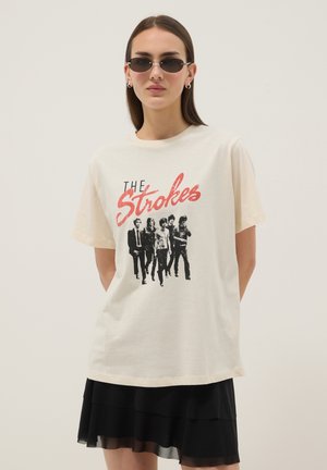 Vrouw met een zonnebril, een crèmekleurig T-shirt met "The Strokes" en een bandfoto, gecombineerd met een zwarte gelaagde rok, staand voor een eenvoudige achtergrond.