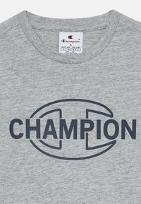 Grå bomull t-skjorte med en stor mørk blå "CHAMPION"-logo i sentrum, med et avrundet emblemdesign som omgir teksten.