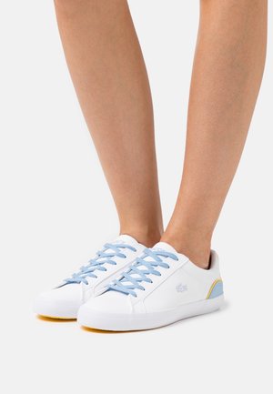 White sneakers with light blue laces, yellow and blue accents. Smooth leather texture, rounded toe, and Lacoste logo on the side.