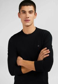 Pull en maille noire à manches longues, col rond et un petit logo sur la poitrine. Le mannequin se tient les bras croisés.