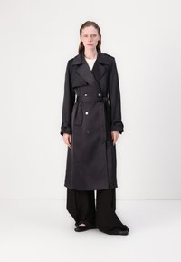 Cappotto grigio con bottoni doppi, ampio colletto, cintura e polsini risvoltati, abbinato a pantaloni neri a gamba larga e scarpe basse.