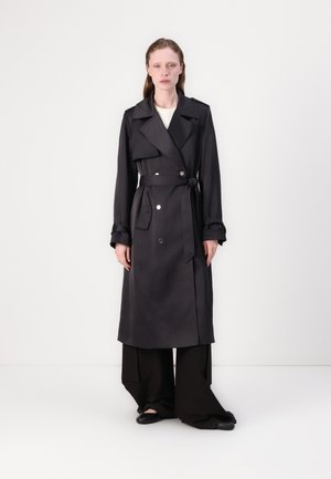 Cappotto grigio con bottoni doppi, ampio colletto, cintura e polsini risvoltati, abbinato a pantaloni neri a gamba larga e scarpe basse.