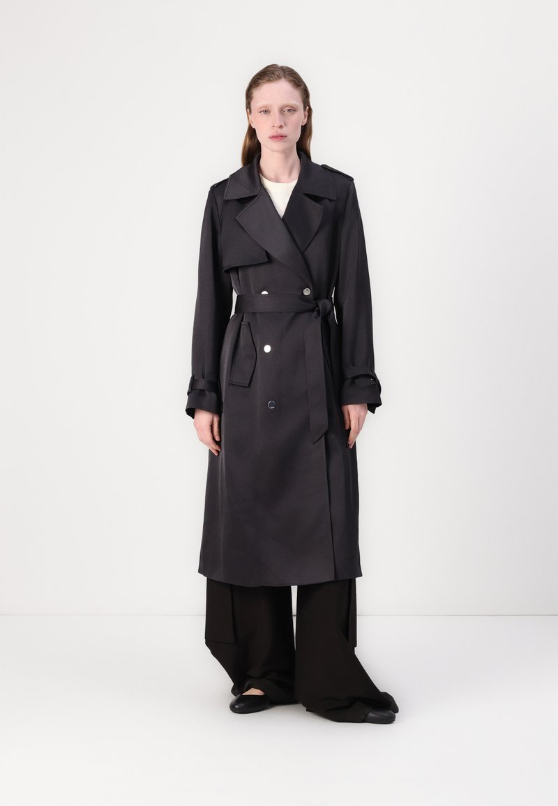 Cappotto grigio con bottoni doppi, ampio colletto, cintura e polsini risvoltati, abbinato a pantaloni neri a gamba larga e scarpe basse.