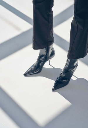 Une personne portant un pantalon noir et des bottines brillantes à talons hauts et à bout pointu se tient en plein soleil, projetant des ombres sur un sol clair.