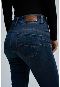 Jeans de mezclilla azul oscuro con un diseño ajustado, cintura alta y costuras en contraste de color naranja. Presenta dos bolsillos traseros y una etiqueta de cuero marrón.