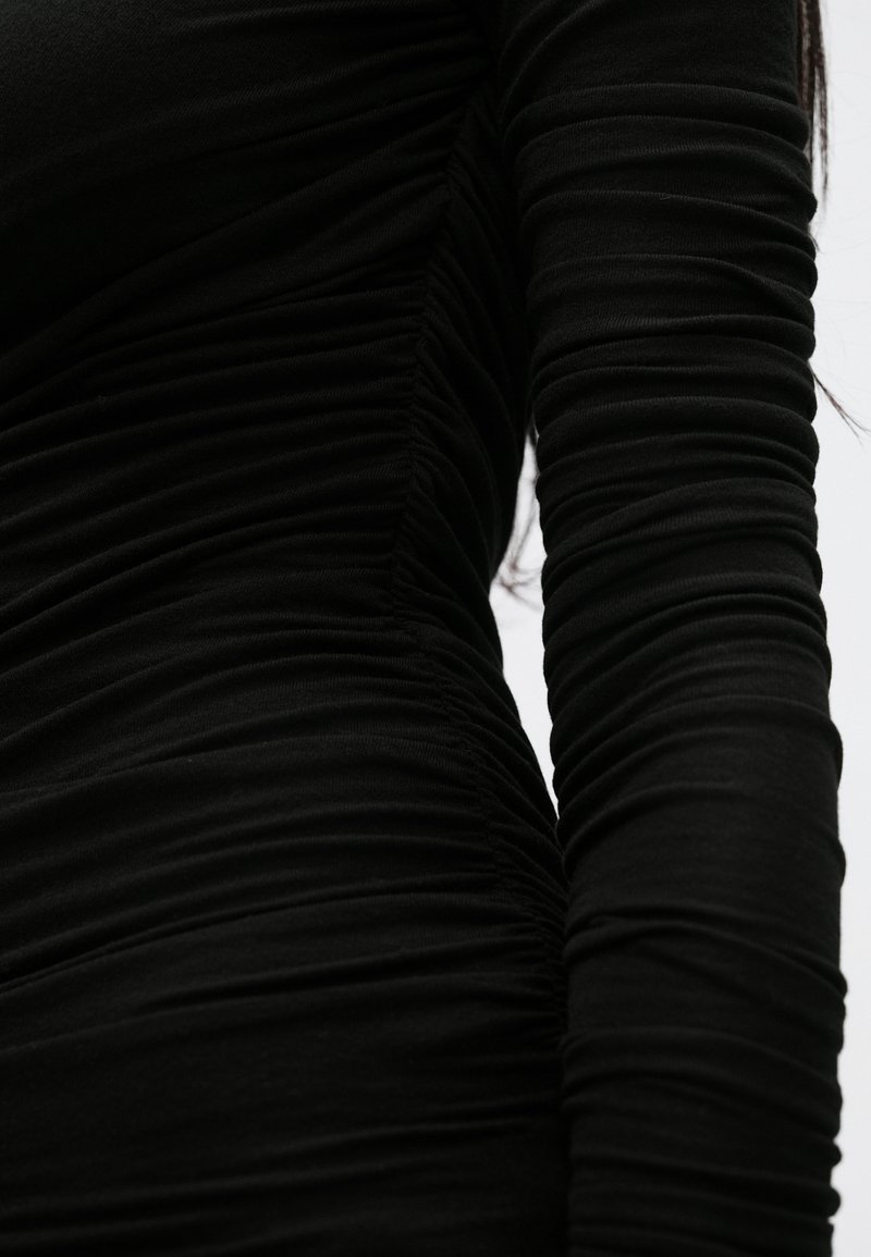 Schwarzes Langarmkleid mit strukturiertem Rüschendesign, das entlang der Seiten und Ärmel gerafften Stoff für eine figurbetonte Silhouette aufweist.