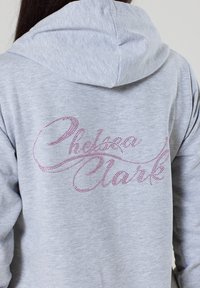Grauer Kapuzenpullover mit einem Strassdesign auf dem Rücken, das "Chelsea Clark" in einer geschwungenen Schriftart zeigt. Glatter Stofftextur.