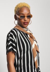 Óculos de sol retangulares castanhos, blusa listrada em preto e branco, lenço de cetim castanho claro amarrado no pescoço, brincos grandes em forma de argola, cabelo texturizado.