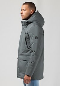 Pilkas pūkinis švarkas su kapišonu, su užtrauktuku užsegamu priekiniu kišeniu ir dideliu priekiniu kišeniu. Lygus paviršius, lengvas dizainas.