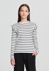 Camicia a maniche lunghe bianca con righe orizzontali nere, con maniche arricciate e scollatura a girocollo. Indossata con pantaloni neri.