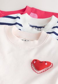 Kolm kokku volditud laste särk roosas, valges siniste triipudega ja ühtlases punases toonis, roosal särgil naeratava südame lapiga.