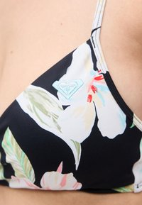 Κοντινό πλάνο μαύρου τοπ μπικίνι με πολύχρωμο φλοράλ σχέδιο και ανοιχτό γαλάζιο κεντημένο λογότυπο στο στήθος, που φοριέται από ένα άτομο.