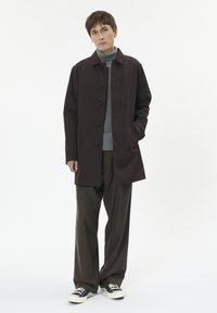 Brązowy płaszcz zapinany na guziki z kołnierzem, długimi rękawami i bocznymi kieszeniami, noszony na szarym swetrze i brązowych spodniach, w połączeniu z czarnymi trampkami.