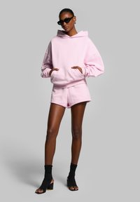 Rosa hoodie och shorts-set i mjukt tyg. Hoodien har en ficka framtill och en snörning i huvan. Svarta öppna tå-klacksandaler.