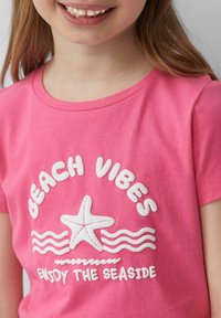 Roze katoenen T-shirt met de tekst "BEACH VIBES" en "ENJOY THE SEASIDE" in het wit, met een sterfiscusgrafiek en golvende lijnen.