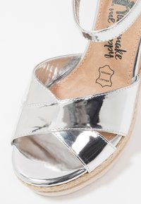 Sandale en cuir métallique argenté avec des brides croisées, une bride à la cheville et une semelle compensée en liège montrant l'étiquette en cuir véritable de la semelle intérieure.