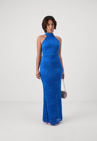 Mėlyna nėriniuota halter tipo suknelė su gėlių raštu, apgauti siluetas ir mermaid tipo apačia. Modelis laiko mažą, raštuotą rankinę.