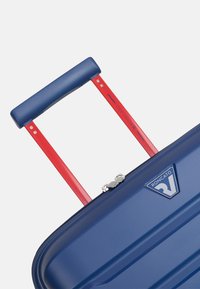 Valigia rigida con superficie blu testurizzata, manico retrattile rosso e zip argentata. Presenta un logo sulla parte anteriore.