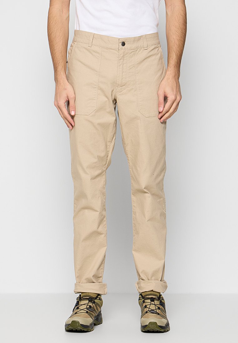 Pantalons en coton beige avec une coupe droite, comportant deux poches avant et une fermeture à bouton. Les ourlets retroussés laissent entrevoir les chaussures.