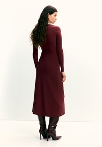 Dunkelrotes, langärmeliges Kleid mit tailliertem Oberteil und ausgestelltem Rock, gefertigt aus glattem Stoff. Kombiniert mit kniehohen, spitz zulaufenden braunen Stiefeln.