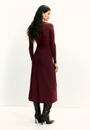 Bordó hosszú ujjú ruha, testhezálló felsőrésszel és bővülő szoknyával, sima anyagból készült. Térdig érő, hegyes orrú barna csizmával párosítva.