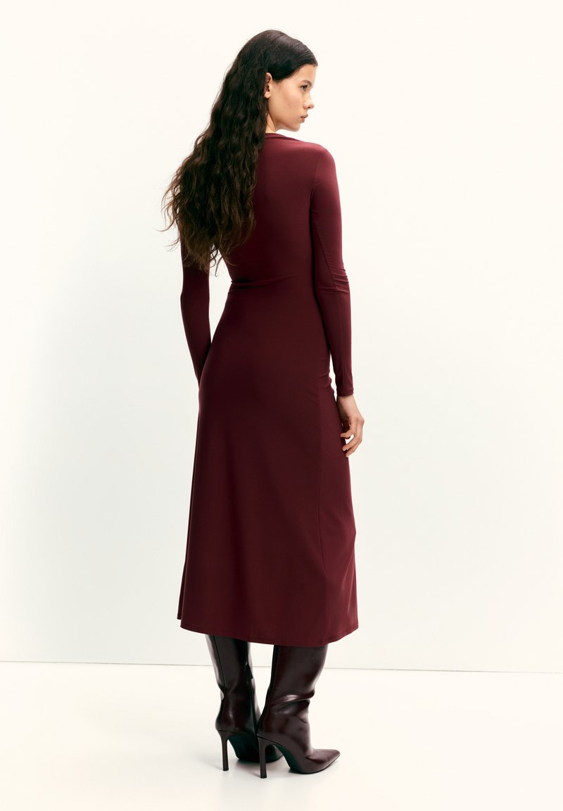 Rochie bordo cu mâneci lungi, corset strâmt și fustă evazată, confecționată dintr-un material neted. Asortată cu cizme maro până la genunchi, cu vârf ascuțit.