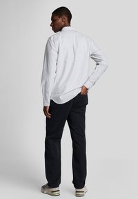 Vit och marinrandig långärmad skjorta med ett veck i ryggen, tillsammans med mörka byxor och grå sneakers, sedd bakifrån.