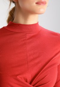 Red cotton top featuring a high neck, smooth texture, and gathered accent detail at the waist. The fabric appears lightweight.