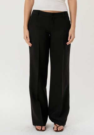 Kobieta nosząca czarne spodnie o szerokich nogawkach i brązowe sandały z odkrytymi palcami, stojąca na jednolitym białym tle.