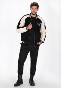 Sort og creme bomberjakke med en fuzzy tekstur, kontrasterende ærmer, stribet krave og broderi foran. Kombineret med sorte bukser og støvler.