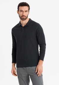 Man med skägg som bär en svart långärmad polo-tröja och grå cargobyxor, stående mot en vit bakgrund.