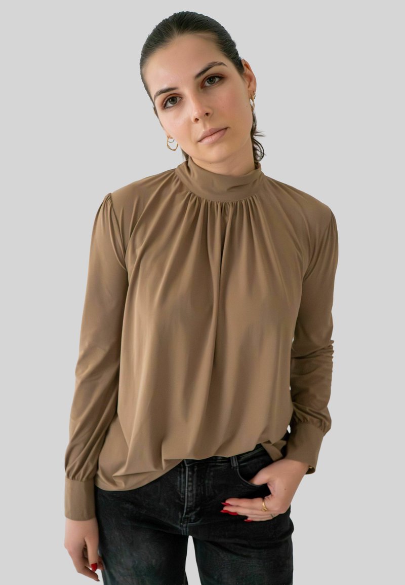 Jonge vrouw met donker haar in een staart, gekleed in een bruine blouse met hoge kraag en zwarte jeans, staand tegen een effen lichtgrijze achtergrond.