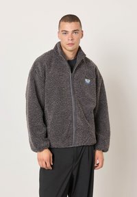 Jaunas vyras su trumpais plaukais, dėvintis pilką, tekstūruotą striukę su užtrauktuku ir juodas kelnes, stovintis priešais paprastą šviesią foną.