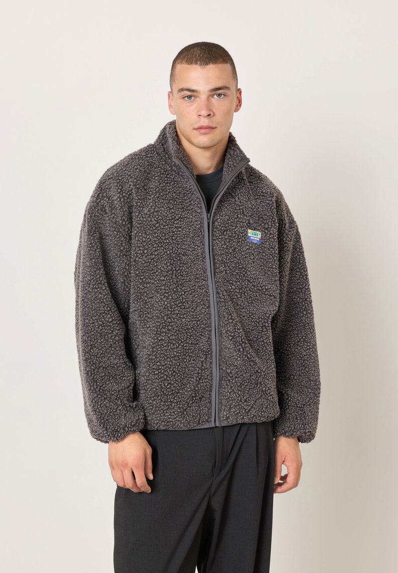 Jaunas vyras su trumpais plaukais, dėvintis pilką, tekstūruotą striukę su užtrauktuku ir juodas kelnes, stovintis priešais paprastą šviesią foną.