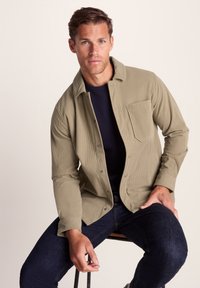 Man with short brown hair wearing a beige button-up corduroy shirt over a black shirt, dark jeans, sitting on a stool against a plain background.