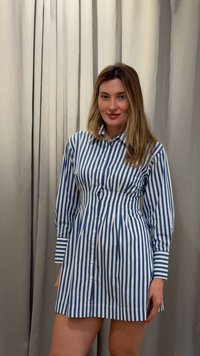 Femeie cu păr lung și blond poartă o rochie cu nasturi, cu dungi verticale albastre și albe, stă în fața unor draperii bej și zâmbește ușor.