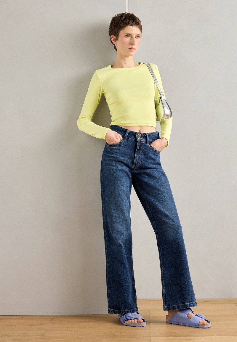 Hellgelbes, langärmeliges, cropped Oberteil, hochtaillierte blaue Jeans und lavendelfarbene Sandalen. Strukturierter Stoff, lässige Passform mit Knopfdetails an der Hose.