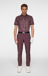 Męski strój golfowy składający się z koszulki polo z krótkim rękawem w kolorze bordowym z białymi wzorami konturowymi oraz dopasowanych bordowych spodni, uzupełniony czarnym paskiem i białymi butami.