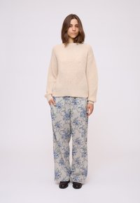 Pull en tricot crème à manches longues, associé à un pantalon large à motif floral gris. Le mannequin porte des chaussures noires, avec une coiffure simple.
