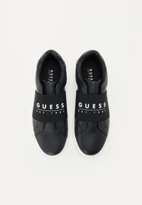 Sneakers in pelle nera con bande elastiche riportanti "GUESS EST. 1981" in bianco. Texture liscia con punta tonda e suole bianche a contrasto.