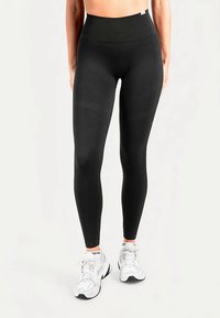 Leggings noirs taille haute fabriqués dans un matériau lisse et extensible. Ils présentent une conception sans coutures, une coupe ajustée et une étiquette de marque minimaliste.