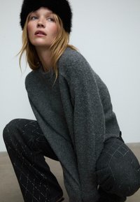Szary sweter z dzianiny o okrągłym dekolcie, zestawiony z ciemnoniebieskimi spodniami dżinsowymi z srebrnymi diamentowymi wzorami. Modelka nosi czapkę z czarnej futrzanej tkaniny.