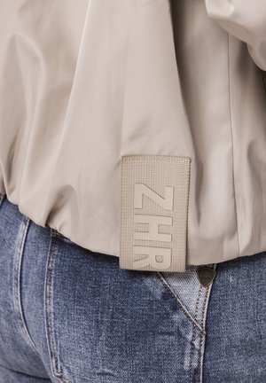 Beige Jacke mit einem rechteckigen gewebten Etikett mit der Aufschrift "ZHR", das über der Tasche einer blauen Jeans mit einem kleinen, dreieckigen Metalllogo hängt.