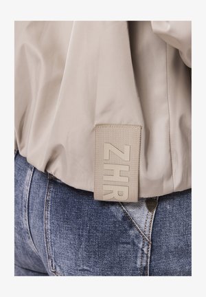 Beige jacket with a rectangular woven label reading "ZHR" hanging over blue denim jeans pocket with a small triangular metal logo.