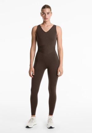Bruine aansluitende jumpsuit met een V-hals en kruisdetaillering bij de taille, gemaakt van een gladde, rekbare stof, gecombineerd met witte sportschoenen.
